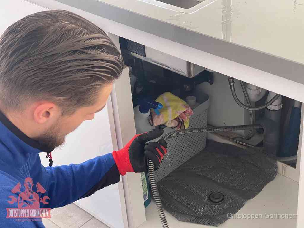 Een monteur voert een flexibele ontstoppingsveer in onder het keukenkastje, langs de sifon, om de gootsteen te ontstoppen in Gorinchem