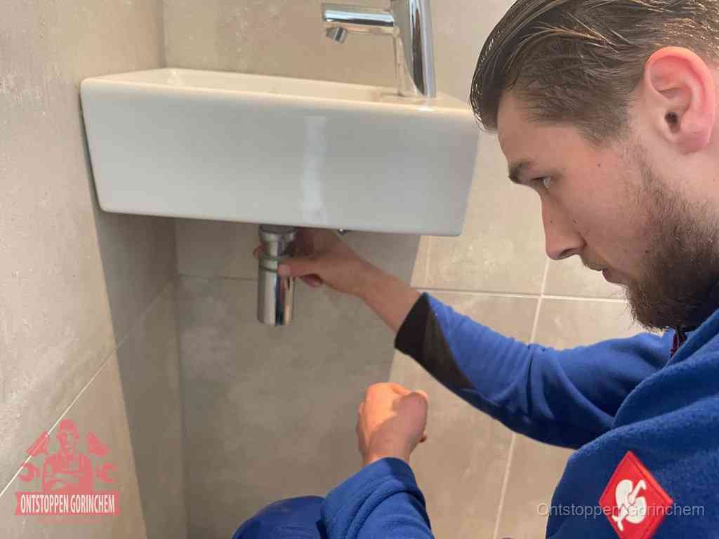 Een monteur draait met de hand de zwanenhalssifon onder een fonteinwasbak in de toiletruimte los om de bron van rioollucht in Gorinchem te vinden